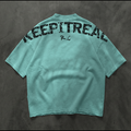 A mint green oversized short-sleeve t-shirt shown from the back, featuring the word 'KEEPITREAL' printed in a distressed black font across the upper back and a small 'R.C' monogram below it, laid flat on a dark textured background.
