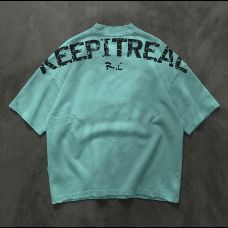 A mint green oversized short-sleeve t-shirt shown from the back, featuring the word 'KEEPITREAL' printed in a distressed black font across the upper back and a small 'R.C' monogram below it, laid flat on a dark textured background.